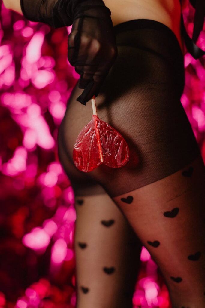 A stylish and glamorous woman holding a heart-shaped lollipop against a pink backdrop.